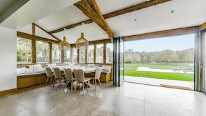 Een ruime eethoek met een grote tafel en stoelen, houten balken in het plafond, en grote ramen met uitzicht op een groen landschap.