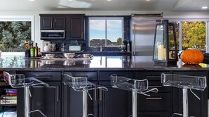 Une cuisine moderne avec des meubles de couleur sombre, un grand îlot central doté de tabourets de bar et une vue sur l’extérieur.