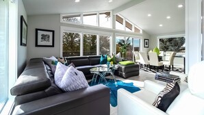 Un vaste espace de vie comprenant un canapé en cuir foncé, une table basse en verre et un canapé blanc agrémenté de coussins à motifs. De nombreuses fenêtres laissent entrer la lumière naturelle et offrent une vue sur l’extérieur.