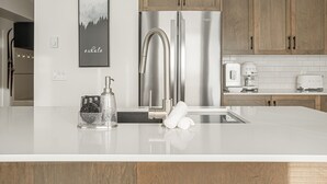 A modern kitchen with a stainless steel refrigerator, wooden cabinets, a white countertop, a faucet, and a coffee maker. - Modern Happy Valley Executive TH with Hot Tub (Beaverdell)