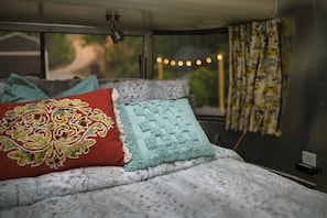 Una cama con ropa de cama estampada y almohadas decorativas, una ventana con cortinas y una guirnalda de luces.