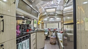 Cocina compacta con microondas, estufa y lavamanos. Hay un banco con un otomano estampado y una pequeña mesa con un cartel que dice "Happy Campers".
