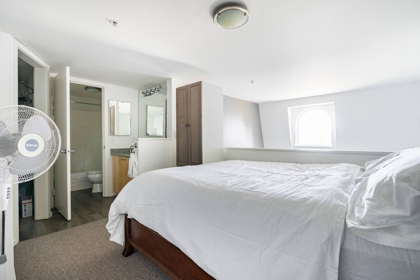 A bedroom with a large bed, a standing fan, a window, and a door leading to a bathroom.