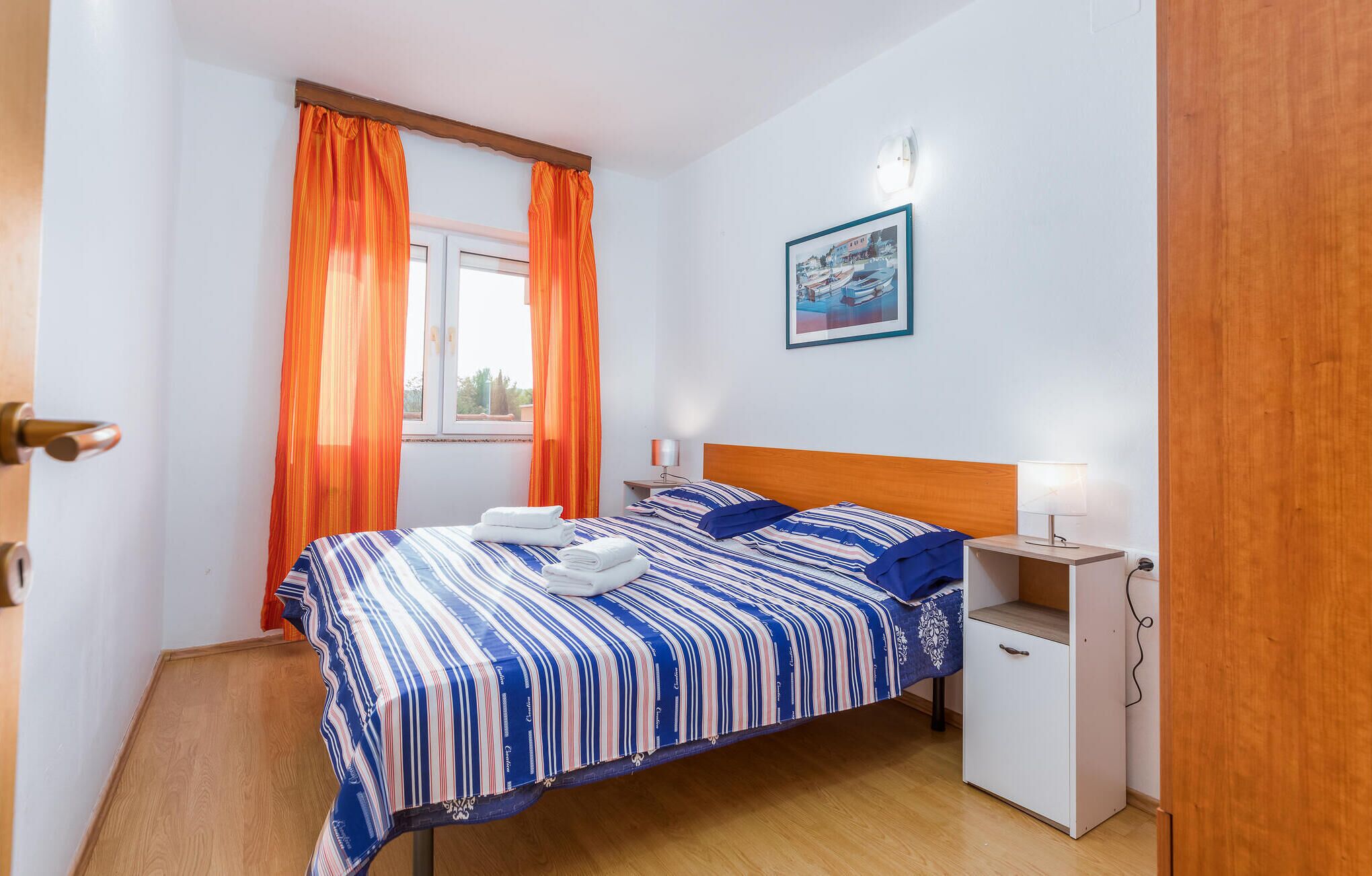 A bedroom with a bed, two bedside tables, a nightstand, a window with curtains, and a wall-mounted picture.