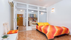 A compact room with a bed, a small table, a chair, and a kitchenette visible through an open door.