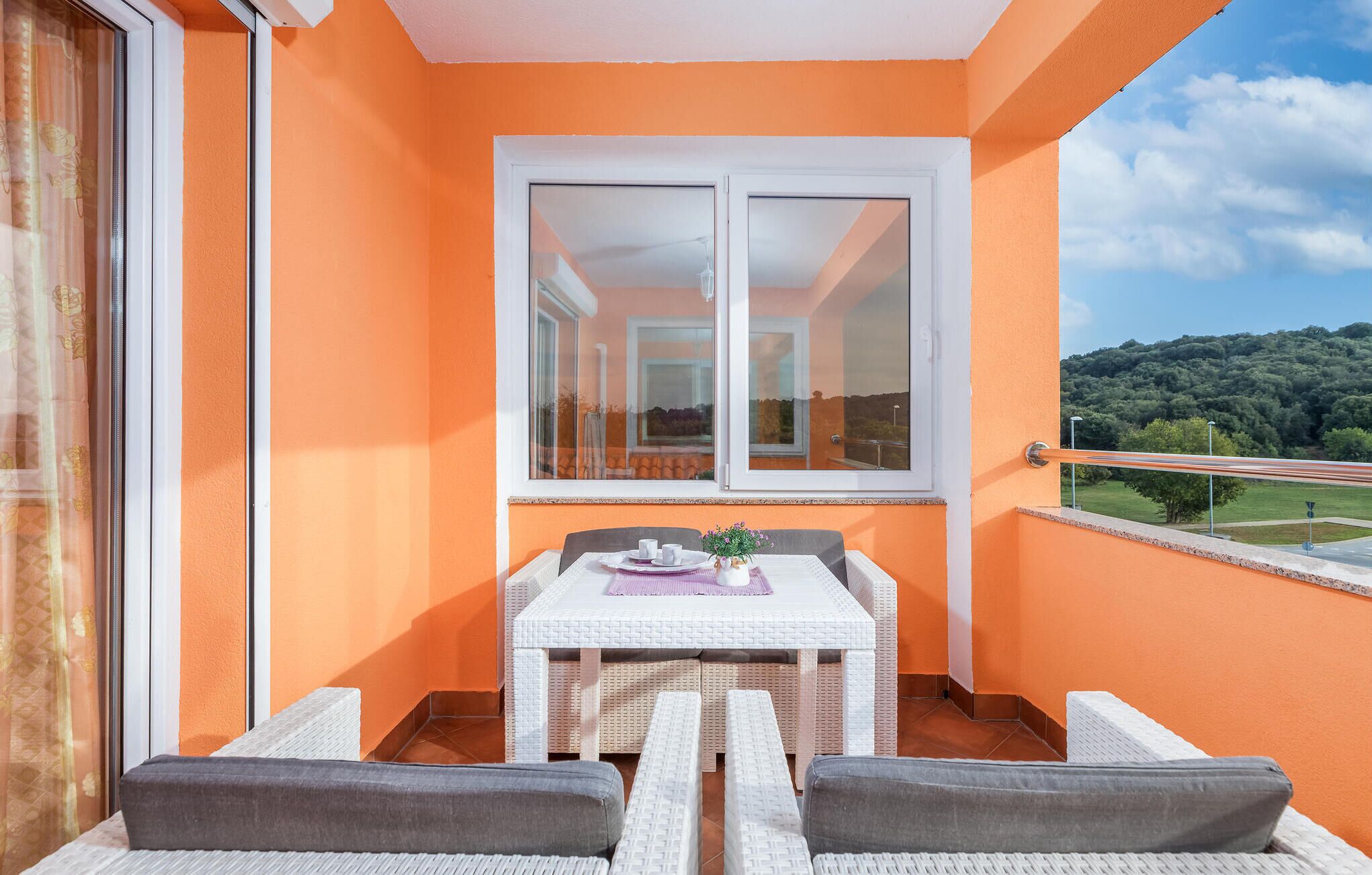 A balcony with a white table and two chairs, a window, and a view of a green landscape.