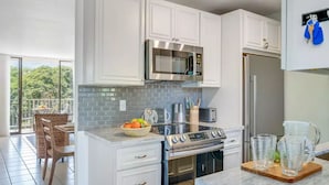 Cocina moderna con armarios blancos, microondas de acero inoxidable y estufa a gas. Hay un comedor con mesa y sillas, y un balcón con vistas a zonas verdes.