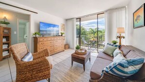 Un salón con sofá, una silla de mimbre, un aparador de madera y un televisor. Hay un balcón con vistas a zonas verdes.