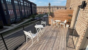 Een houten terras met zitplaatsen buiten, uitzicht op een bakstenen gebouw en een heldere lucht.