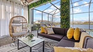 En moderne stue med en fletstol, en sofa med gule puder, et glasbord og udsigt over vand gennem store vinduer.