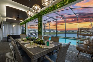 A dining area with a view of a sunset over a body of water, featuring a large table, wicker chairs, and a glass-enclosed pool. - Lakeview Luxury Villa in Storey Lake (Kissimmee)