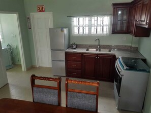 A kitchen with a refrigerator, stove, sink, and wooden cabinets. There is a window with bars and an exit sign on the door.
