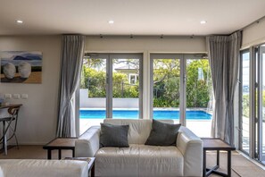 A modern living room with a pool view, a sofa, and a dining area. - Hauraki Vista (Waiheke Island)