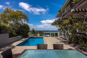 A swimming pool with a view of the ocean and hills, surrounded by greenery and a modern building. - Hauraki Vista (Waiheke Island)