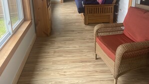 Een gezellige kamer met een houten vloer, een blauwe bank met gestreepte kussens, een rieten stoel met een rode kussen, en een grote houten deur.