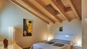Un dormitorio con techo de madera, una cama con colcha gris y beige, dos mesitas de noche con lámparas y un cuadro en la pared.