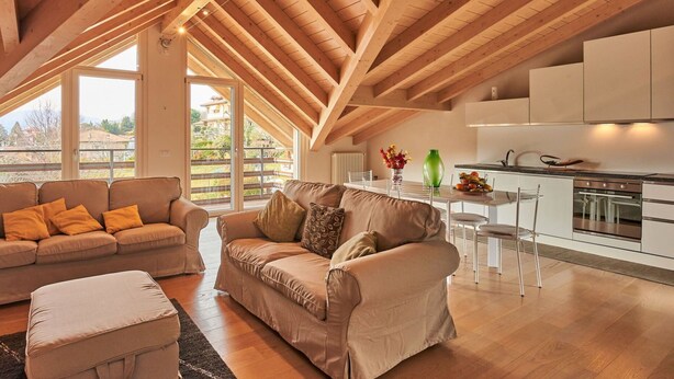 Un amplio salón con techo alto y vigas de madera, una cocina moderna con armarios blancos y vistas a un jardín a través de grandes ventanales.