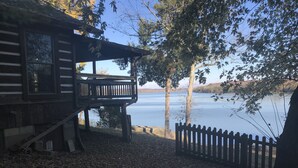 A cabin with a deck overlooking a lake, surrounded by trees and a wooden fence. - Derby Time Riverside Cabin (Derby)