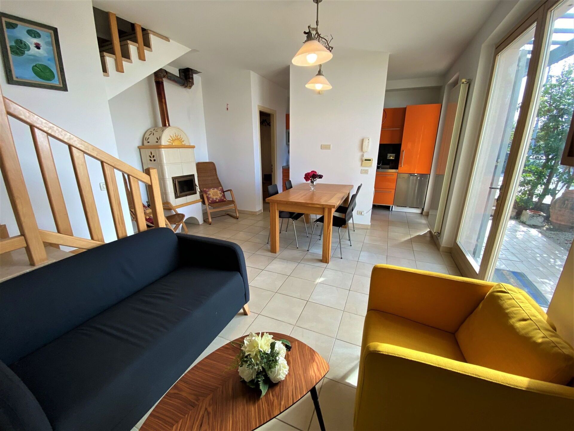 A living room with a sofa, a yellow armchair, a wooden coffee table with a flower, a dining table with chairs, a fireplace, and a kitchen in the background.