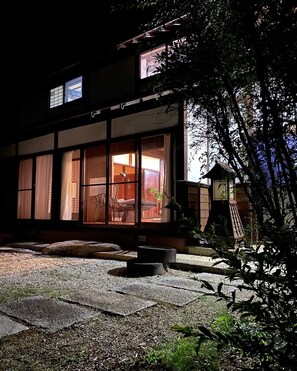Ein traditionelles japanisches Gebäude mit großen Schiebegläsern, einem gepflasterten Innenhof und einer kleinen Holzhütte im Hintergrund.