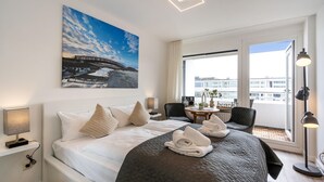 A hotel room with a bed, two chairs, a small table, and a large window offering a view of buildings.