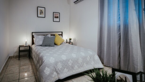 Een slaapkamer met een bed, nachtkastje, airconditioner en een raam met gordijnen.