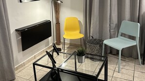 Een woonkamer met een flatscreen televisie, een ingelijst kunstwerk, een gele stoel, een lichtblauwe stoel, een glazen salontafel en een vloerlamp.