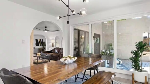 A modern dining area with a wooden table, chairs, and a bowl of fruit. There is a view of a patio with plants and a ceiling fan. - 6 bd Desert Oasis w/ Pool, Slide, Pickleball+More! (Scottsdale)