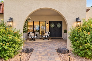 An outdoor patio area with a brick floor, two striped armchairs, a small table, and a dark door with decorative wreaths.