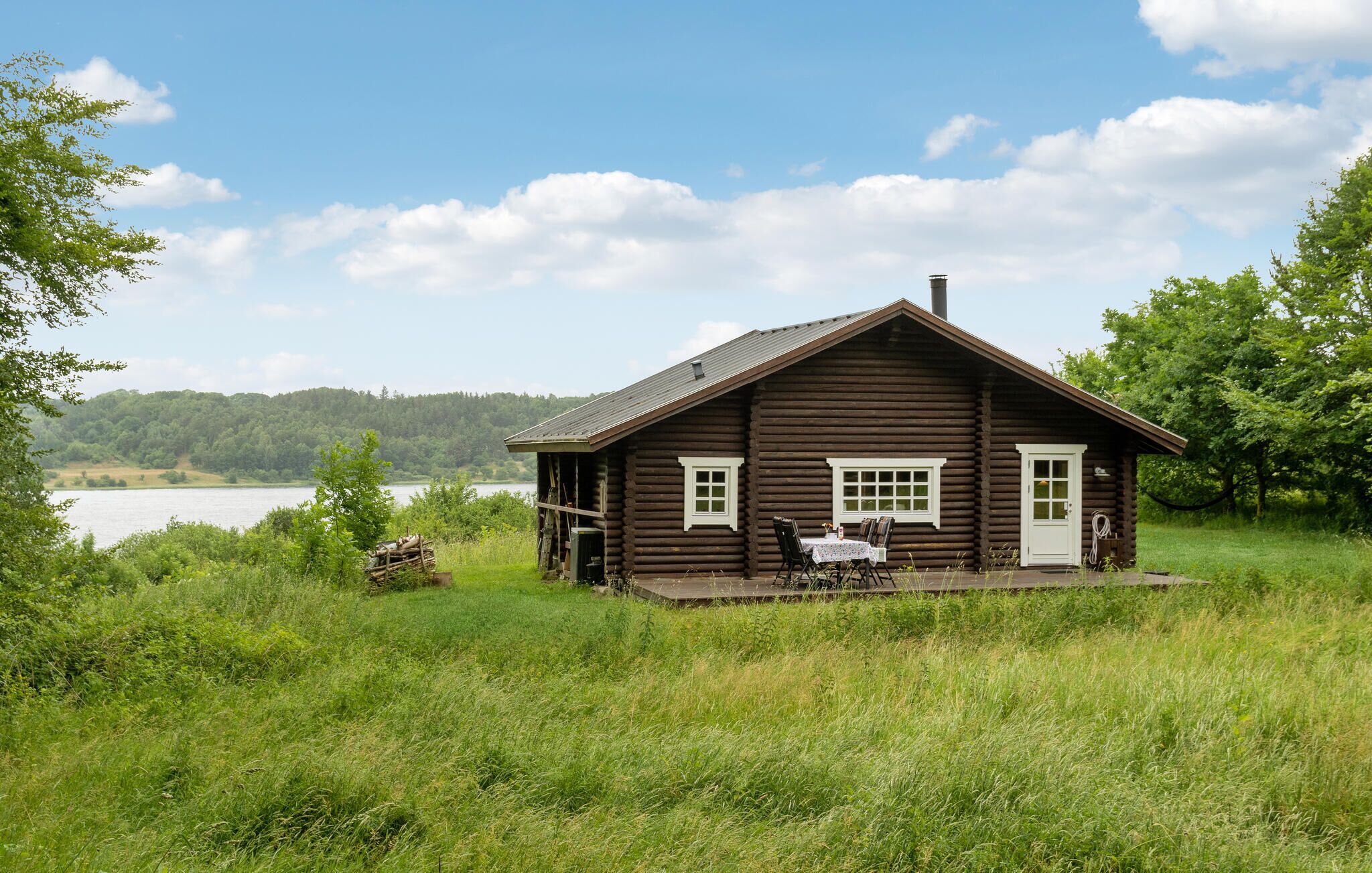 Een knus houten huisje met een veranda, een tafel en stoelen, en uitzicht op een meer en bos.