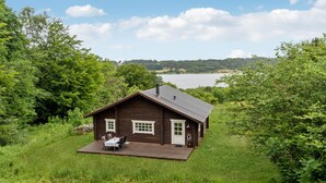 A wooden cabin with a deck, surrounded by lush greenery and a lake in the background. - 2 bedroom cozy home in Mariager (Mariager)