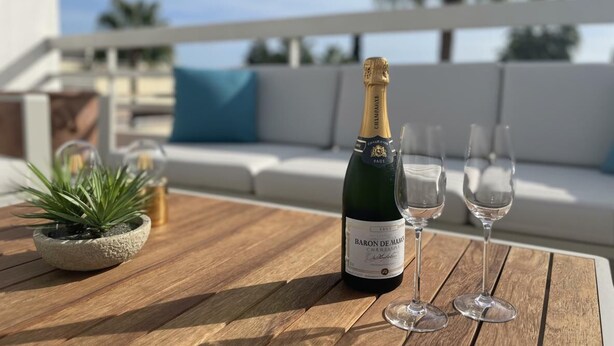 A bottle of Baron de Champagne on a wooden table with two empty wine glasses, a small potted plant, and a white couch with a blue cushion in the background. - Pent-House, 3 Bedroom, Sleeps 6, Parking (Marbella)