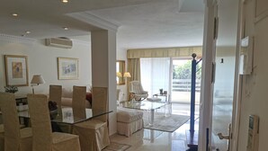 A modern living room with a glass coffee table, beige armchairs, and a sofa. There is a dining area with a glass table and chairs. The room has large windows with curtains, a view of greenery outside. - Pent-House, 3 Bedroom, Sleeps 6, Parking (Marbella)