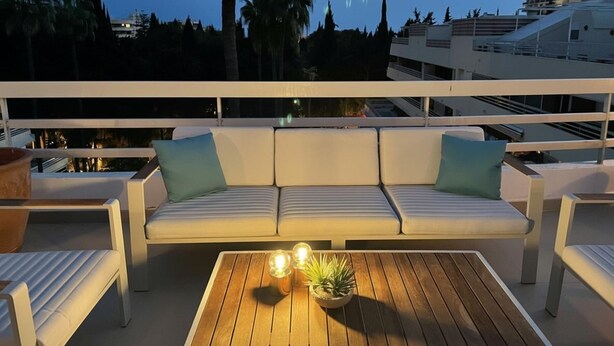 En balkon med en hvid sofa, to puder, et træbord med en plante og to stearinlys.