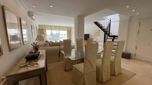 A spacious living area with a glass-top dining table, beige upholstered chairs, a sofa, and a staircase leading to an upper floor. - Pent-House, 3 Bedroom, Sleeps 6, Parking (Marbella)