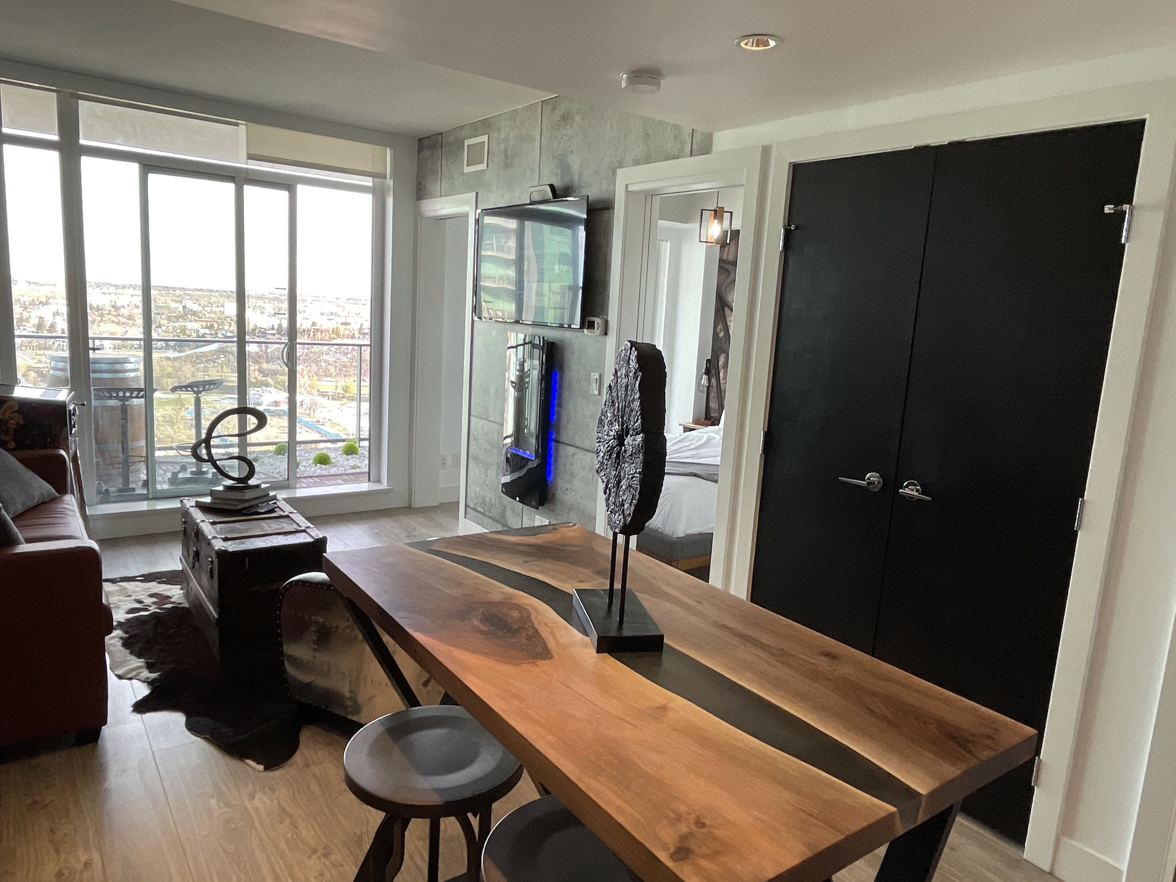 A modern kitchen with a wooden table, bar stools, a large window, and a black wardrobe.