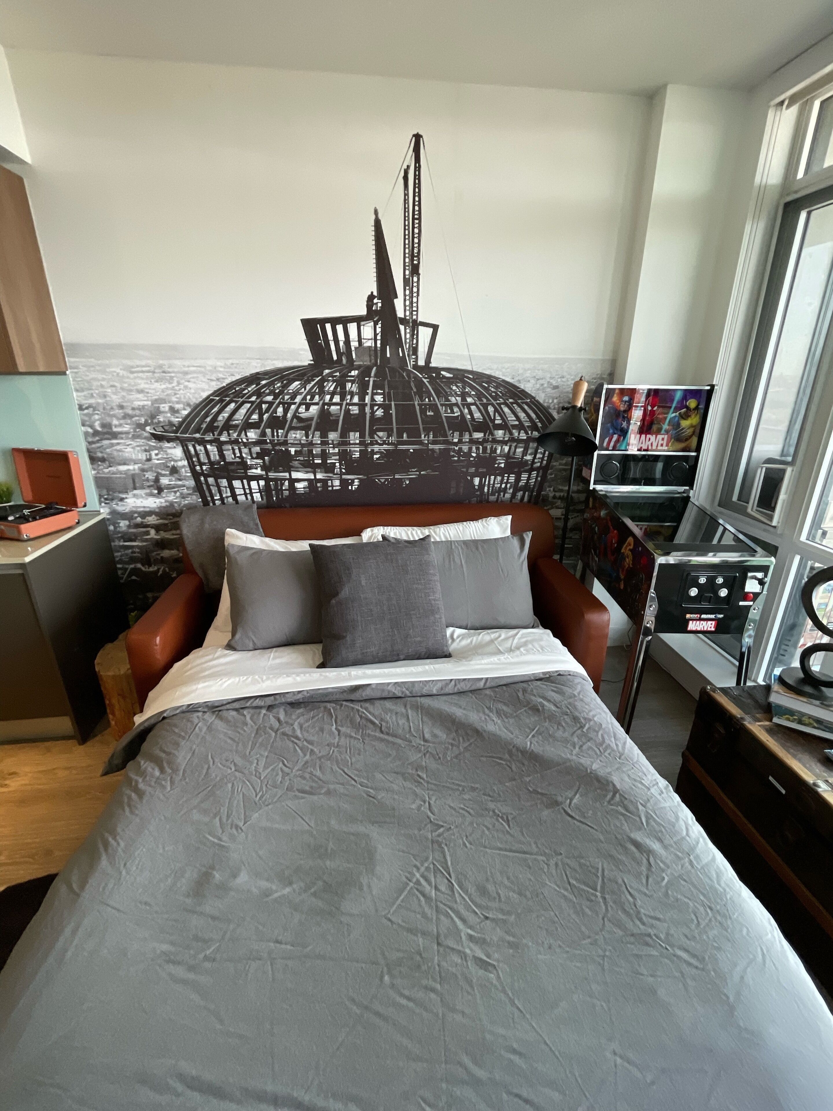 A bedroom with a bed, a headboard with a cityscape design, a desk with a computer, and a window.