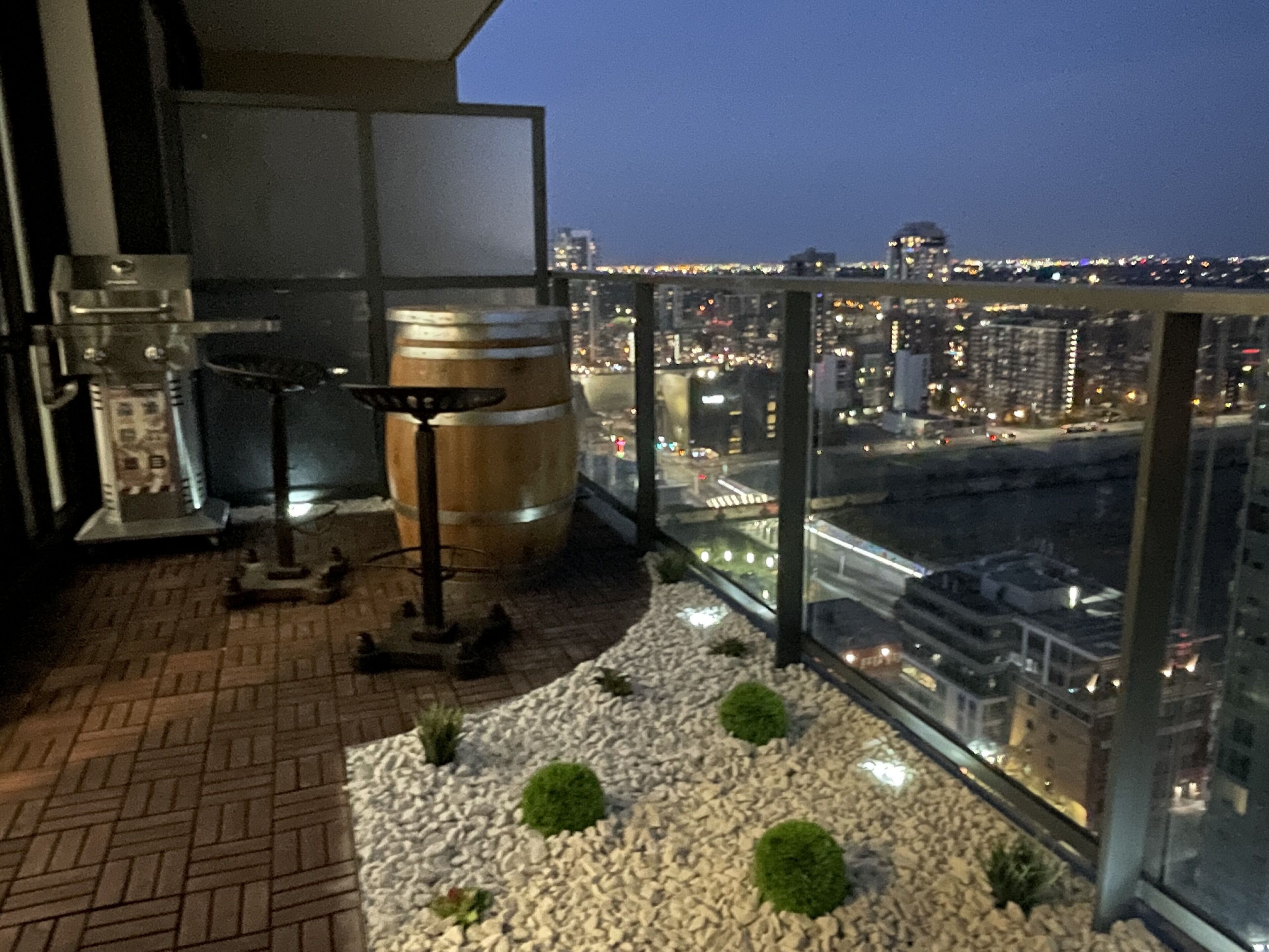 Un balcone con vista sulla città di notte, con una grande botte di metallo, un tavolino e una pianta in vaso.
