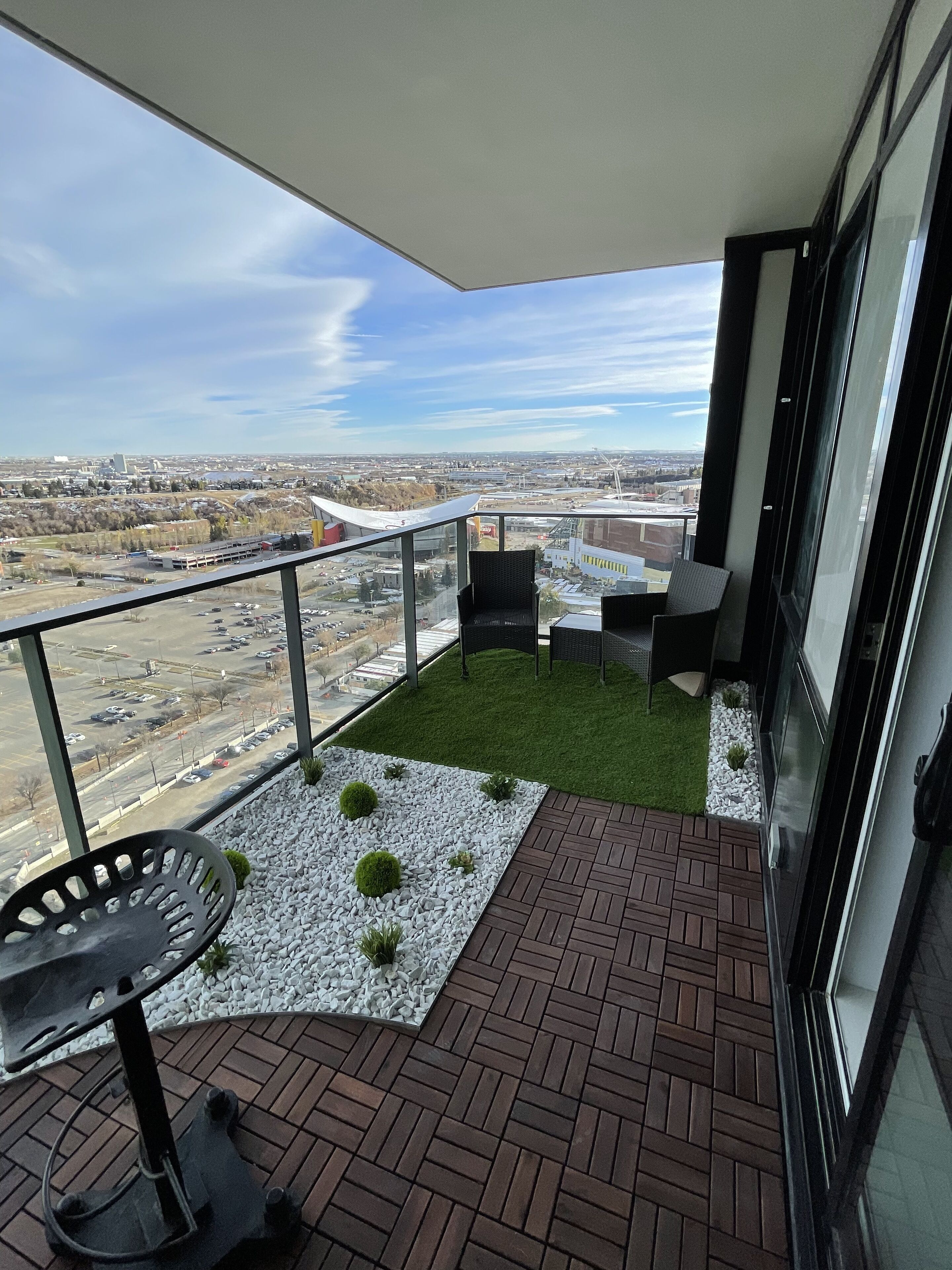 A balcony with a view of a cityscape, a small seating area with a table and chairs, and a grassy patch with decorative plants.