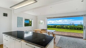 A modern kitchen with a large island, a dining area with a table and chairs, and a sliding glass door leading to a patio with a view of the ocean. - Palm Ridge - near Onetangi & Palm Beach (Waiheke Island)