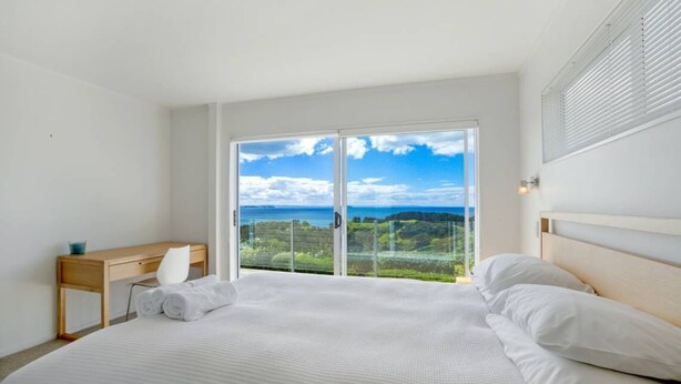 A bedroom with a large bed, a wooden desk, and a view of the outdoors through a sliding glass door. - Palm Ridge - near Onetangi & Palm Beach (Waiheke Island)