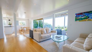 A spacious living room with a large window, a sofa, a glass coffee table, and a dining area with a table and chairs. - Palm Ridge - near Onetangi & Palm Beach (Waiheke Island)