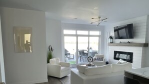 Ein modernes Wohnzimmer mit einem weißen Sofa, einem Couchtisch und einem an der Wand befestigten Fernseher. Ein großes Fenster sorgt für natürliches Licht und bietet einen Blick ins Freie.