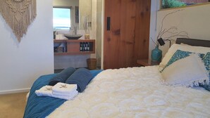 Een slaapkamer met een bed, een houten garderobe, een badkamer met een wastafel en spiegel, en een macramé wanddecoratie.
