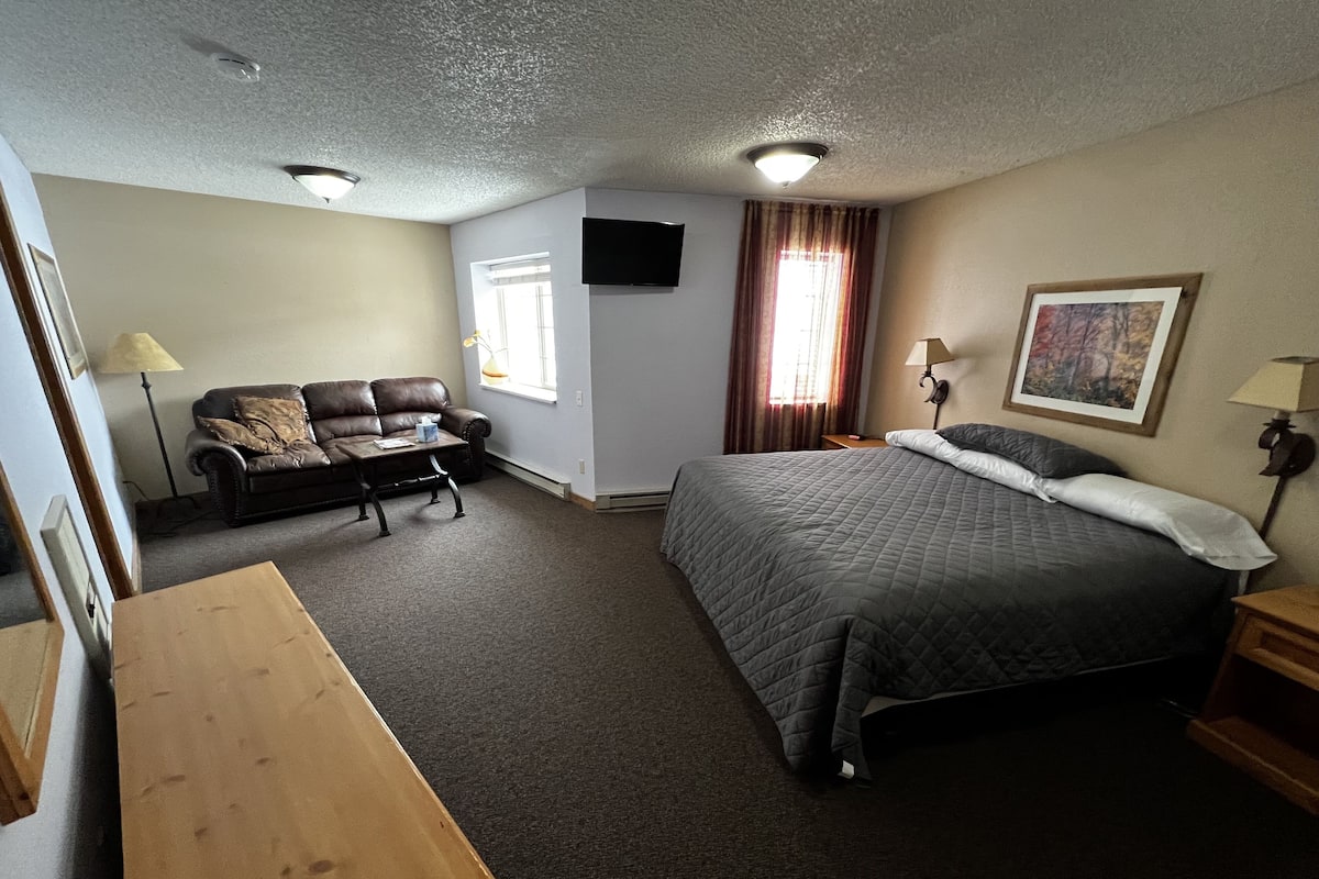 A hotel room with a bed, a sofa, a desk, a flat-screen TV mounted on the wall, and a window with curtains.