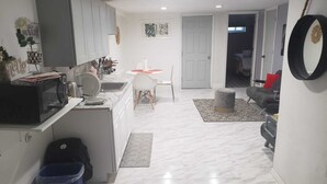 Una cocina moderna con armarios blancos, un microondas y un lavamanos. Hay una zona de comedor con una mesa y sillas blancas, y un pequeño otomaní. El piso está cubierto de baldosas con diseño de mármol.