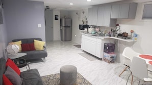 Una sala de estar moderna con un sofá gris, un tapete estampado y un reposapiés gris. Se ve una cocina al fondo con armarios blancos y un refrigerador de acero inoxidable.