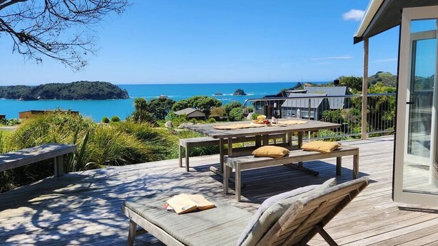 Eine Holzterrasse mit Tisch und Bänken, die auf eine Küstenlandschaft mit klarem, blauem Wasser und weiter Landpartie blickt.
