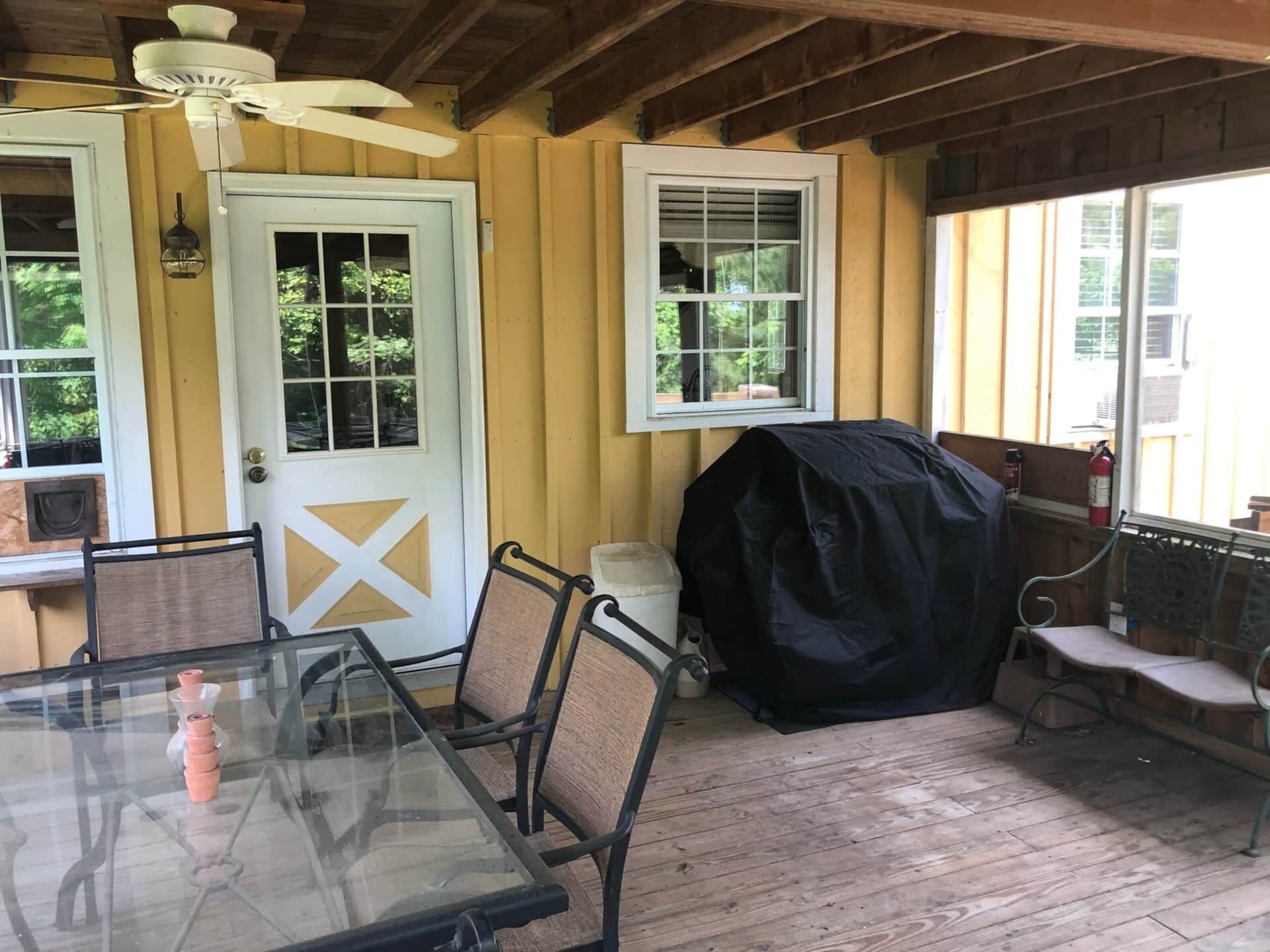 Un patio techado con mesa de vidrio, sillas, una parrilla y un ventilador de techo.