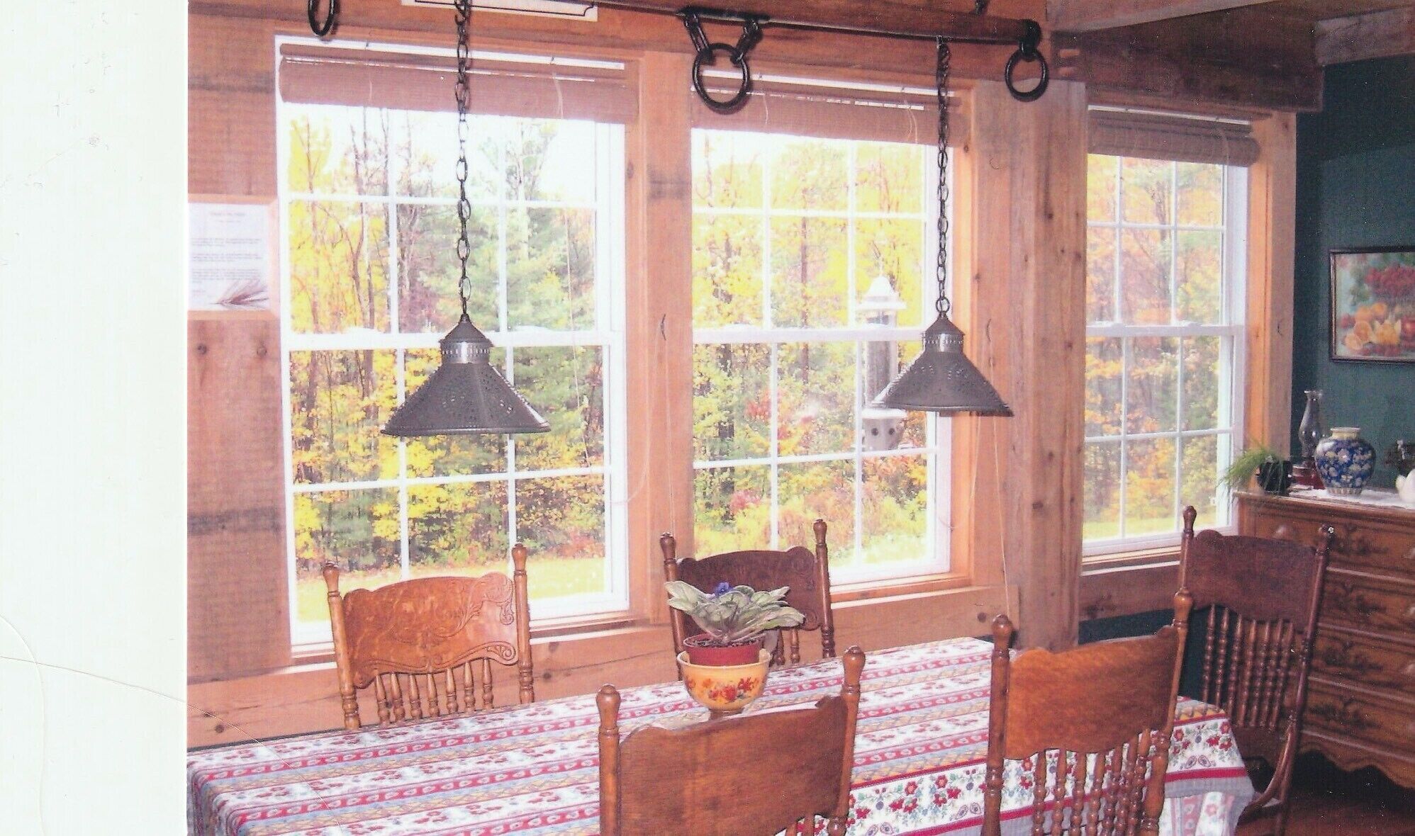 A rustic dining room with wooden chairs, a table with a patterned tablecloth, hanging lamps, and large windows with a view of autumn trees.
